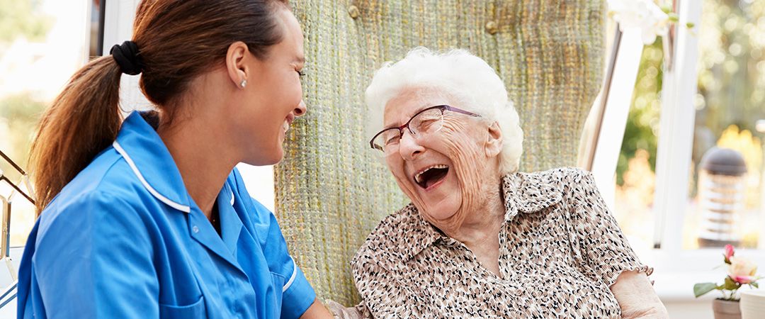 Healthcare worker in blue scrubs sitting outside with senior woman who is laughing.